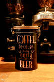Schwarze Kaffeedose mit Bambusdeckel und Bügelverschluss in Kaffeebar, vor zwei Kaffeeautomaten, rosegoldener Spruch: COFFEE because it´s