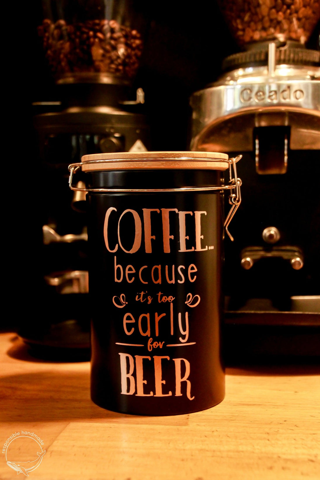 Schwarze Kaffeedose mit Bambusdeckel und Bügelverschluss in Kaffeebar, vor zwei Kaffeeautomaten, rosegoldener Spruch: COFFEE because it´s