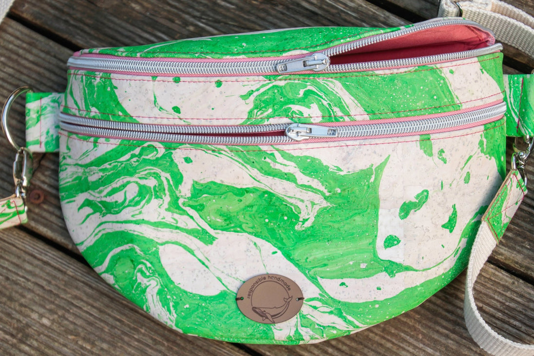 Slingbag aus handmarmoriertem Kork, Farbe grün und weiß mit zwei Reißverschlussfächern, weißem Gurtband auf Holzhintergrund